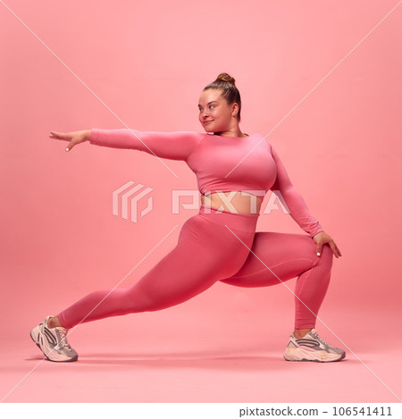 Full body portrait of young plus size sporty woman in pink top and legging training, stretching legs and hands isolated studio background. 106541411