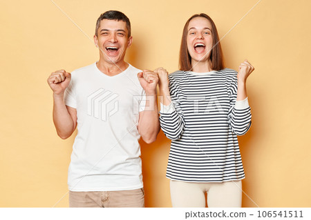 Extremely happy young couple friends man woman in casual clothing isolated over beige background clenched fists yelling happily celebrating their achievement. Extremely happy young couple friends man woman in casual clothing isolated over beige background clenched fists yelling happily celebrating their achievement. 106541511