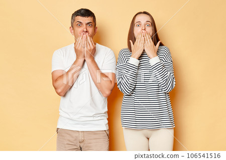 Shocked astonished young couple wife and husband wearing casual attires standing isolated over beige background looking at camera with big eyes covering mouths with hands. 106541516