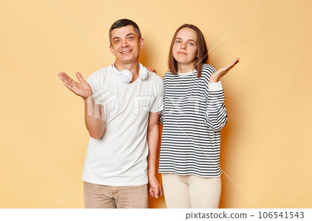 Puzzled young couple wife and husband wearing casual attires standing isolated over beige background shrugging shoulders saying they can't help looking at camera. Puzzled young couple wife and husband wearing casual attires standing isolated over beige background shrugging shoulders saying they can't help looking at camera. 106541543
