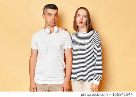 Funny childish young couple wife and husband wearing casual attires standing isolated over beige background frimacing showing tongue out and pout lips playful behavior. Funny childish young couple wife and husband wearing casual attires standing isolated over beige background frimacing showing tongue out and pout lips playful behavior. 106541544
