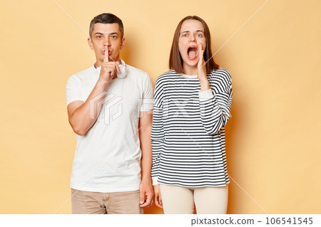 Caucasian woman and man wearing casual outfits standing isolated over beige background, guy showing shh gesture, female screaming loud with hand near mouth. 106541545