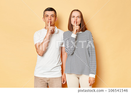 Serious strict young couple friends man woman in casual clothing isolated over beige background keeping fingers near mouths keep secret looking at cmaera with bossy expressions. Serious strict young couple friends man woman in casual clothing isolated over beige background keeping fingers near mouths keep secret looking at cmaera with bossy expressions. 106541546