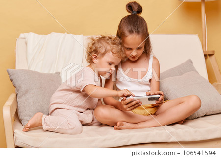 Concentrated children little girls sisters sitting on couch using smartphone together posing against beige wall playing on mobile phone looking at device display. Concentrated children little girls sisters sitting on couch using smartphone together posing against beige wall playing on mobile phone looking at device display. 106541594
