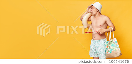 Astonished man wearing shorts swimsuit and panama holding donut rubber ring isolated on yellow background looking far to advertisement area copy space for promotional text. 106541676