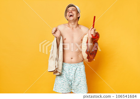 Extremely happy positive adult man wearing shorts swimsuit and panama holding donut rubber ring and bag isolated on yellow background rejoicing resting on tropical resort. 106541684