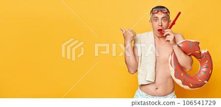 Surprised astonished man in diving goggles and snorkel holding donut rubber ring isolated on yellow background pointing away presenting area for advertisement. 106541729
