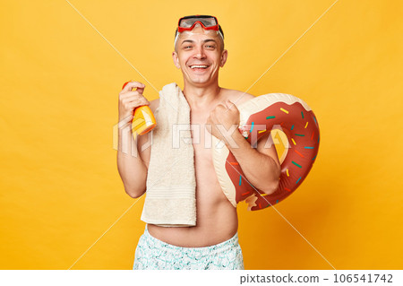 Happy satisfied adult man wearing shorts swimsuit snorkeling goggles holding donut rubber ring and bag isolated on yellow background holding sunscreen for skin care during sunbathing. 106541742