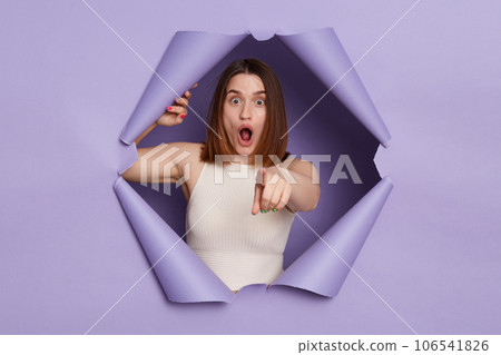 Shocked surprised brunette woman breaking through purple paper hole wearing casual white top pointing at camera choosing you keeps mouth open. 106541826