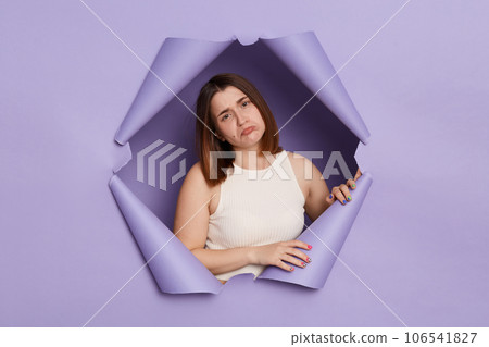 Sad upset bored brunette woman breaking through purple paper hole wearing casual white top looking at camera with pout lips being in bad mood feeling sorrow. 106541827
