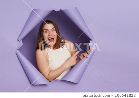 Positive delighted excited brunette woman breaking through purple paper hole wearing casual white top pointing away at copy space for promotion advertisement area. Positive delighted excited brunette woman breaking through purple paper hole wearing casual white top pointing away at copy space for promotion advertisement area. 106541828