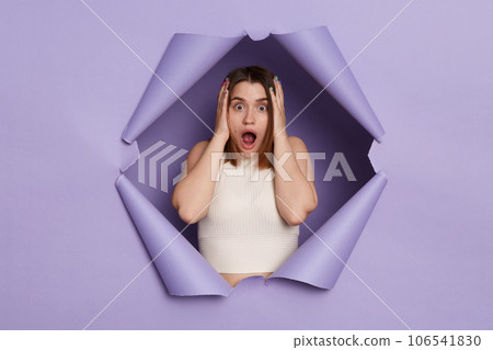 Shocked scared brunette woman breaking through purple paper hole wearing casual white top keeps hand on head sees something frightened saying omg. Shocked scared brunette woman breaking through purple paper hole wearing casual white top keeps hand on head sees something frightened saying omg. 106541830