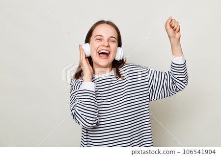 Joyful cheerful young woman wearing striped shirt listening to music in headphones isolated over gray background singing and dancing with raised hand. Joyful cheerful young woman wearing striped shirt listening to music in headphones isolated over gray background singing and dancing with raised hand. 106541904