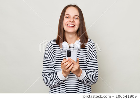 Smiling happy brown haired adult woman wearing striped shirt standing isolated over gray background using smart phone chatting with friends looking at camera and laughing. Smiling happy brown haired adult woman wearing striped shirt standing isolated over gray background using smart phone chatting with friends looking at camera and laughing. 106541909