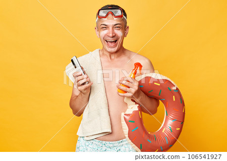 Happy cheerful man in diving goggles and snorkel holding donut rubber ring isolated on yellow background standing with sun protection scream laughing happily. 106541927