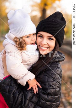 Mother and young little girl with blonde hair are playing in an autumn park on a yellow and orange leaf background Mother and young little girl with blonde hair are playing in an autumn park on a yellow and orange leaf background 106543567