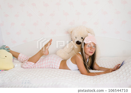 Beautiful teenage girl in pajamas is lying on a cozy bed on her stomach and surfing on the phone in the house in the morning, the younger generation is addicted to gadgets Beautiful teenage girl in pajamas is lying on a cozy bed on her stomach and surfing on the phone in the house in the morning, the younger generation is addicted to gadgets 106544710