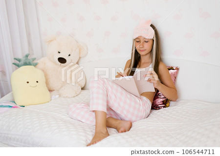 Cute Caucasian teen girl reads her diary sitting on the bed in sleeping clothes and makes notes in it Cute Caucasian teen girl reads her diary sitting on the bed in sleeping clothes and makes notes in it 106544711