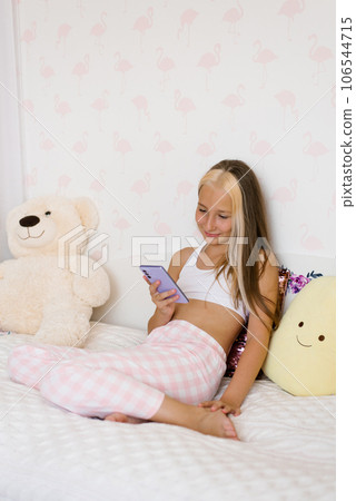Candid shot of cute teenage girl in pajamas sitting on bed, posting pictures on social media and checking friends feed 106544715