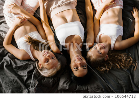 Cheerful family of a smiling mother with two daughters lying together on a bed with dark linens, smiling, looking at the camera, feels happy. The concept of love, offspring, a close-up view from 106544718