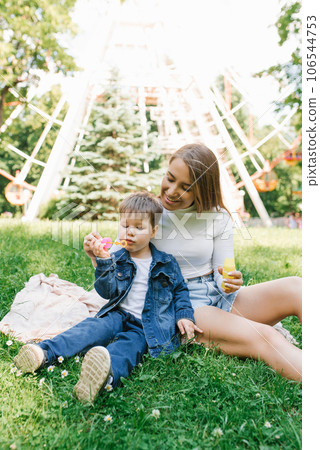 Beautiful cute young mom with her cute little son having fun sitting on the grass in the park outdoors and blowing soap bubbles 106544753
