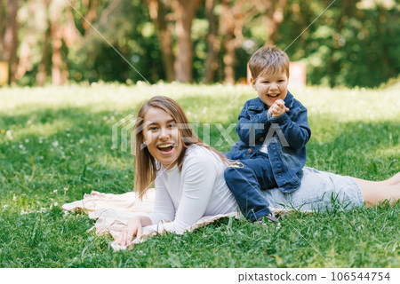 Young Caucasian mother and her young son lie on a blanket on the grass in the park in summer and relax outdoors Young Caucasian mother and her young son lie on a blanket on the grass in the park in summer and relax outdoors 106544754