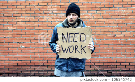 Homeless holds piece of cardboard with inscription Need Work. Refugee is at brick wall looking for job. Male tramp in dirty clothes. Below poverty line. Bearded fugitive beggar. Immigrant. Drunkenness Homeless holds piece of cardboard with inscription Need Work. Refugee is at brick wall looking for job. Male tramp in dirty clothes. Below poverty line. Bearded fugitive beggar. Immigrant. Drunkenness 106545743