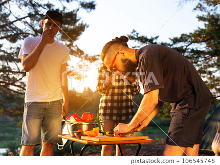 Stylish man with a beard and black glasses cuts fresh vegetables peppers and tomatoes to prepare a salad in nature. Vacation with friends in nature. Tourism as a hobby. Evening sunset. 106545748