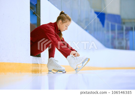 Beautiful girl in red sportswear preparing for training session on ice rink arena, figure skater tying laces on skates Beautiful girl in red sportswear preparing for training session on ice rink arena, figure skater tying laces on skates 106546385