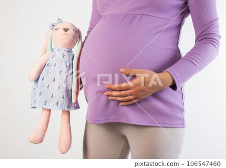 A pregnant girl in a purple blouse holds a soft toy next to her stomach. Concept of motherhood, new life. In anticipation of the birth of the baby. 106547460