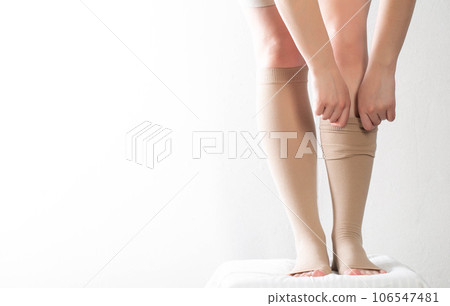 A woman puts on compression stockings for legs with varicose veins. Pain and swelling in the legs, phlebology. White background, close-up. Copy space for text A woman puts on compression stockings for legs with varicose veins. Pain and swelling in the legs, phlebology. White background, close-up. Copy space for text 106547481