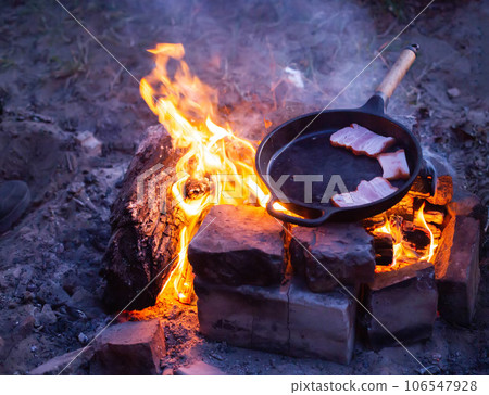 Frying bacon in a pan over a fire. Cooking outdoors in the evening. Frying bacon in a pan over a fire. Cooking outdoors in the evening. 106547928