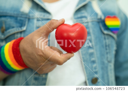Asian lady wearing rainbow flag wristbands and hold red heart, symbol of LGBT pride month celebrate annual in June social of gay, lesbian, bisexual, transgender, human rights. 106548723