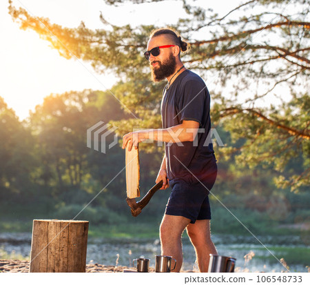 A man with a beard, a lumberjack with an ax in his hands, cuts wood in nature in the summer against the backdrop of a forest and a river bank. Camping. Survival, travel 106548733