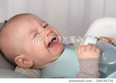 A crying naughty baby holds a bottle of milk formula in his hands and looks at the camera. Baby feeding concept. Copy space for text 106548756