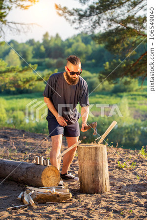 A brutal caucasian man with a beard and an ax in his hands is chopping wood for a fire at a campsite. Outdoor recreation in summer on the banks of the river. Bushcrafting. Sunset 106549040