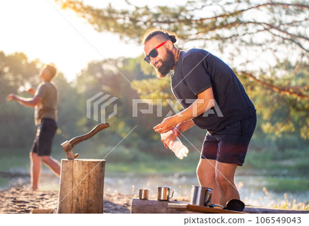 A handsome bearded man in black glasses is chopping firewood in nature in the summer against the background of a sunset. Outing for a picnic. Camping. 106549043