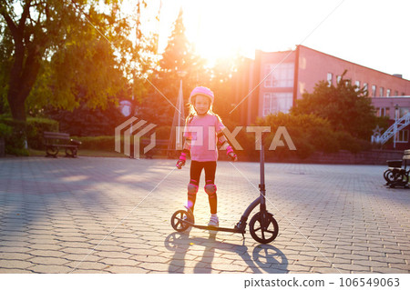 A little caucasian girl in a pink protective gear laughs and grimaces near a scooter against the backdrop of a summer sunset. A little caucasian girl in a pink protective gear laughs and grimaces near a scooter against the backdrop of a summer sunset. 106549063