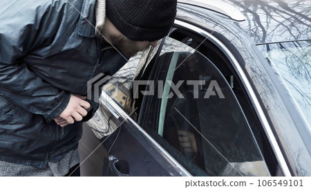 Homeless thief steals purse valuable items from car with unlocked doors open. Property insurance against unlawful theft or crime. Male burglar looks through crack in window. Auto without alarm system 106549101