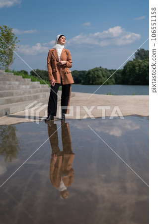 A young woman wearing a hijab stands next to a large puddle. A young woman wearing a hijab stands next to a large puddle. 106551158