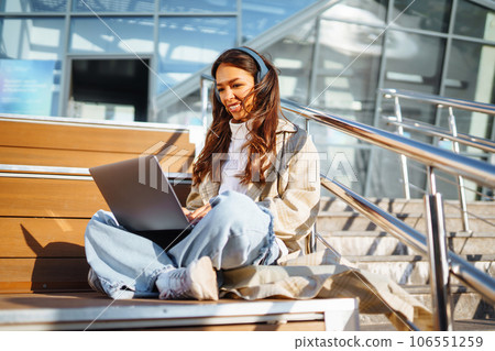 Young woman in headphones with laptop on city street. Video conference. ?nline training. Freelancer Young woman in headphones with laptop on city street. Video conference. ?nline training. Freelancer 106551259