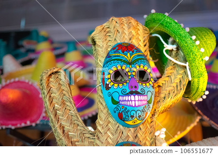 Straw cactus with Mexican blue skull mask and background of colored hats Straw cactus with Mexican blue skull mask and background of colored hats 106551677