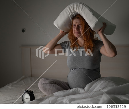 Caucasian pregnant woman sits in bed, holds a pillow on her head and cries. Insomnia. 106552529
