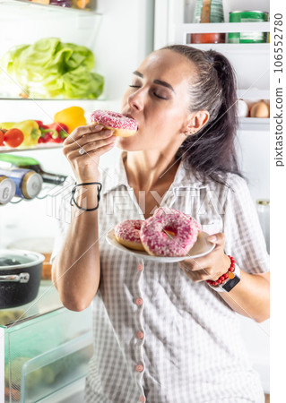 Hungry brunette in pajamas enjoys sweet donuts late at night by the open refrigerator 106552780