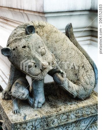 A stone statue of a cat and a kitten. The statue is on a pedestal with a floral design. The cat is in a sitting position with its head turned to the left. The statue is in front of a white wall. 106552883
