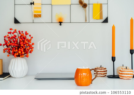 Home office desk with laptop, pumpkin shaped cup with hot drink and marshmallows, seasonal decor like candles, flowers, pumpkins. Autumn inspiration, cozy mood. Hygge home fall decor. Selective focus. Home office desk with laptop, pumpkin shaped cup with hot drink and marshmallows, seasonal decor like candles, flowers, pumpkins. Autumn inspiration, cozy mood. Hygge home fall decor. Selective focus. 106553110