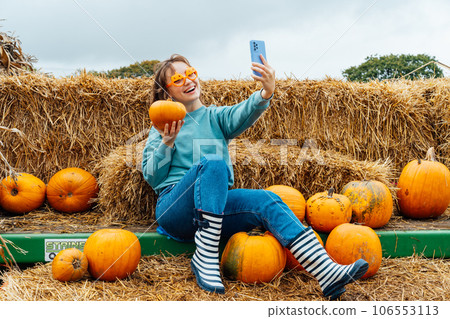 Smiling woman in fun glasses sitting on straw bales and holding pumpkin and making selfie on phone. Selecting Thanksgiving and Halloween holidays decor on agriculture farm. Autumn fall festive mood 106553113