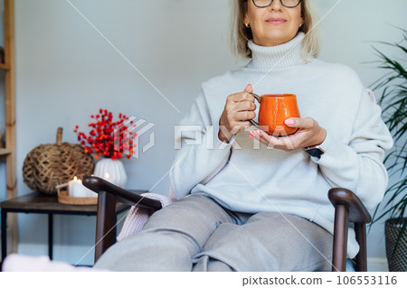 Cropped middle aged woman relaxing with cup of hot drink in scandy style hygge interior home with fall mood decor. Lady dreaming, enjoy calm mood without stress, wellbeing alone. Cozy autumn at home. Cropped middle aged woman relaxing with cup of hot drink in scandy style hygge interior home with fall mood decor. Lady dreaming, enjoy calm mood without stress, wellbeing alone. Cozy autumn at home. 106553116