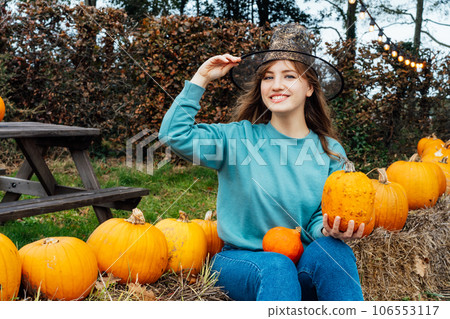 Smiling emotional young woman in witch hat holding pumpkins. Female posing while selecting best pumpkins for Thanksgiving and Halloween holidays decor on agriculture farm. Autumn fall festive mood. Smiling emotional young woman in witch hat holding pumpkins. Female posing while selecting best pumpkins for Thanksgiving and Halloween holidays decor on agriculture farm. Autumn fall festive mood. 106553117