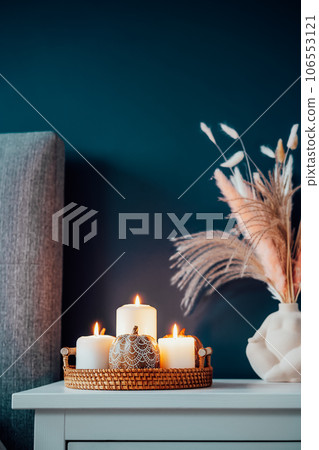 Closeup Autumn decor in stylish modern bedroom. Cozy interior with dark emerald wall, grey fabric headboard, bedside table, vase with pampas grass, pumpkin and burning candles on tray. Vertical card. Closeup Autumn decor in stylish modern bedroom. Cozy interior with dark emerald wall, grey fabric headboard, bedside table, vase with pampas grass, pumpkin and burning candles on tray. Vertical card. 106553121